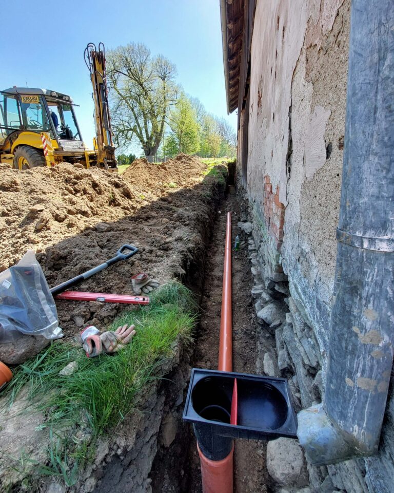 Wąski wykop wzdłuż kamiennej ściany domu; montaż pomarańczowej rury odwodnieniowej z osadnikiem, w tle koparko-ładowarka — wykopy ziemia kłodzka, usługi ziemne Stronie Śląskie.