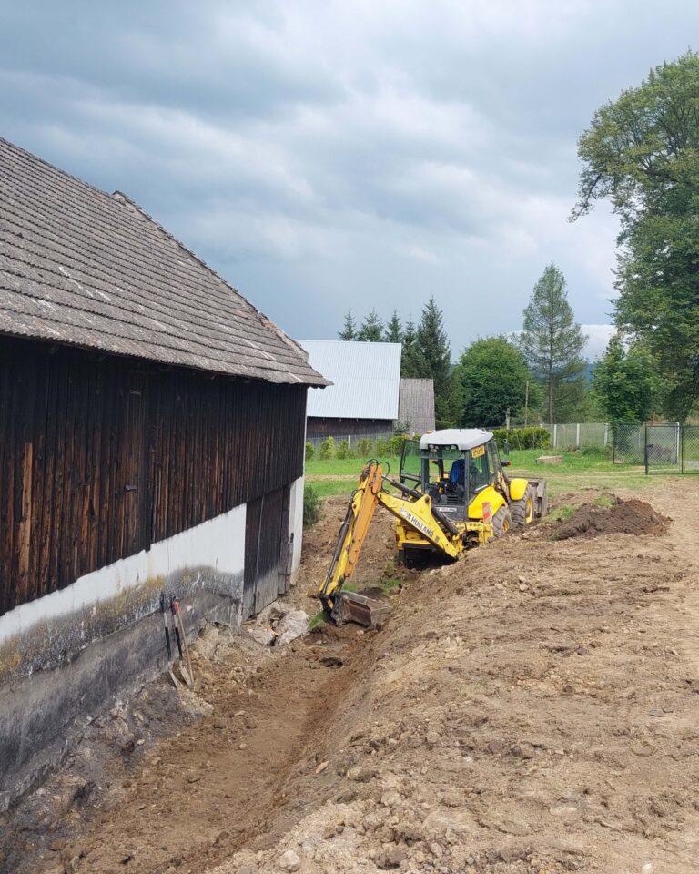 Ramię koparki podsypuje tłuczeń na geowłókninie przy fundamencie; układanie drenażu i podbudowy obok budynku — transport kruszyw ziemia kłodzka, korytowanie Lądek-Zdrój.