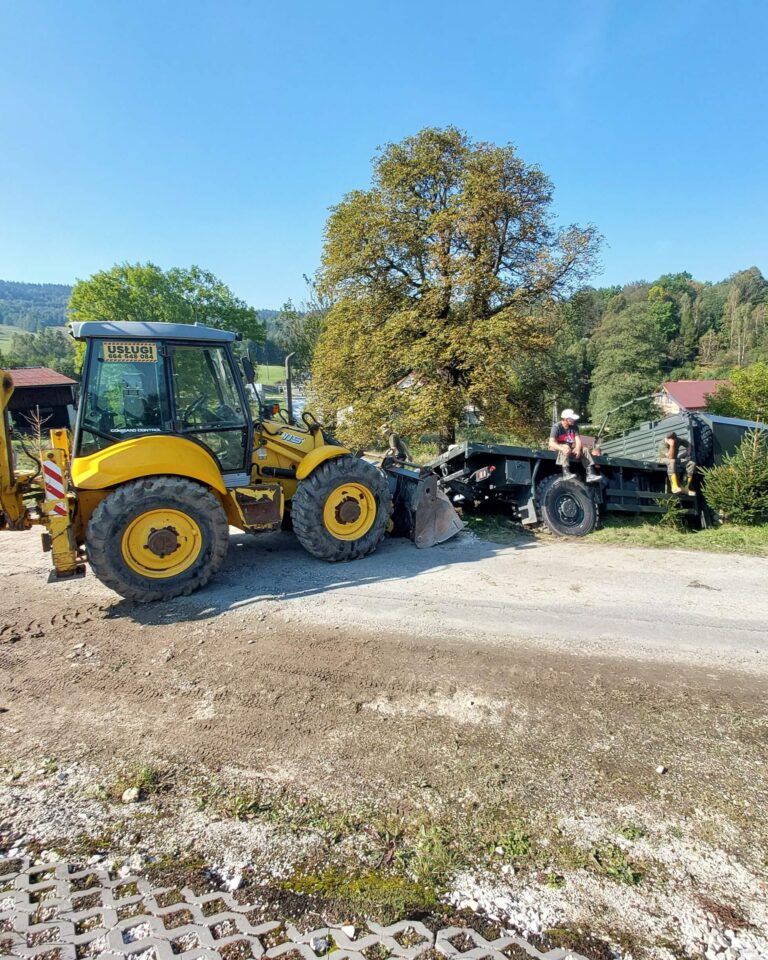 Żółta koparko-ładowarka pomaga podnieść skrzynię wywrotki podczas załadunku na wiejskiej drodze; usługi koparko-ładowarką Ziemia Kłodzka, transport kruszyw Stronie Śląskie.
