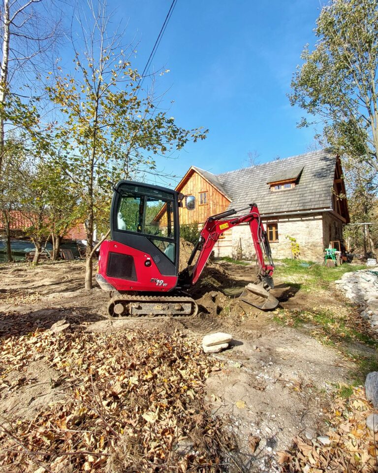 Minikoparka na gąsienicach wyrównuje i korytuje teren obok kamiennego domu, widoczne ślady gąsienic i jesienne liście; roboty ziemne Bystrzyca Kłodzka – korytowanie i utwardzanie terenu.