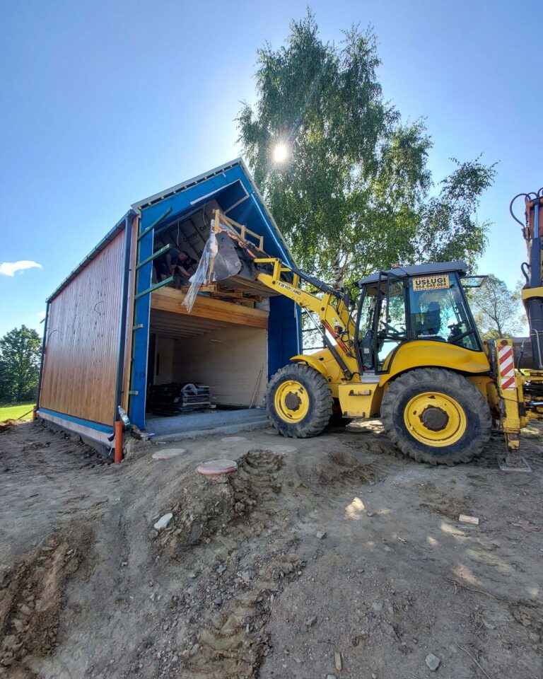 Żółta koparko-ładowarka podnosi paletę z materiałami do nowego domku; słońce nad brzozą, nierówny wjazd – usługi koparko-ładowarką Ziemia Kłodzka, roboty ziemne Stronie Śląskie.