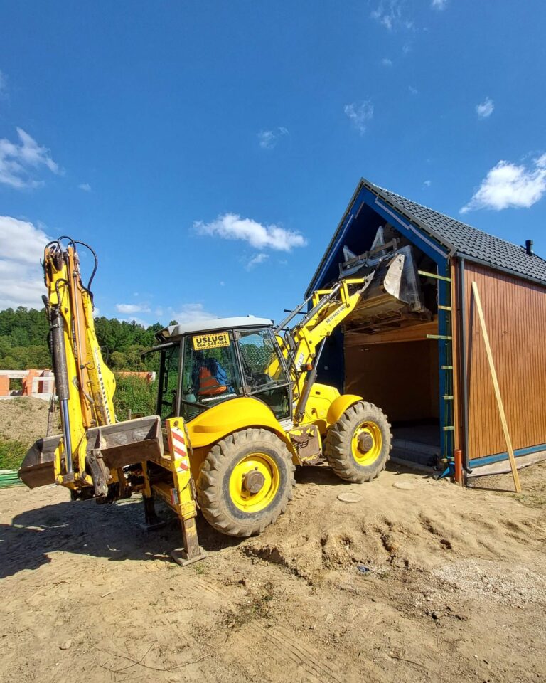 Maszyna z rozłożonymi podporami podaje ładunek przez szczytową bramę drewnianego budynku; plac na wzniesieniu – koparko-ładowarka Kłodzko, usługi ziemne Ziemia Kłodzka.